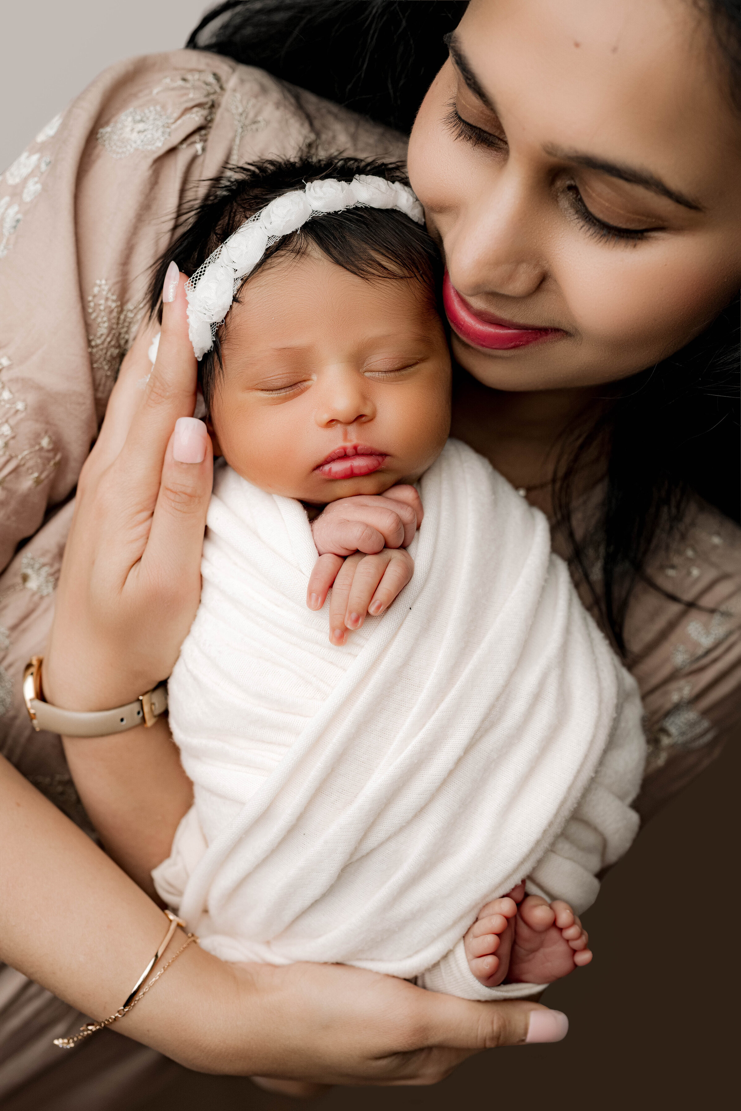 Mother smiles down at her swaddled baby girl, who wears a delicate white wrap and headband.