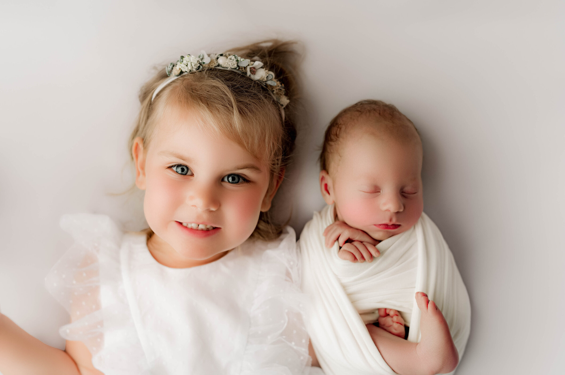 A young girl with a floral headband smiles at the camera, lying next to a sleeping newborn wrapped in a white blanket. Captured by a skilled North Carolina newborn photographer, both rest on a soft, light-colored background.