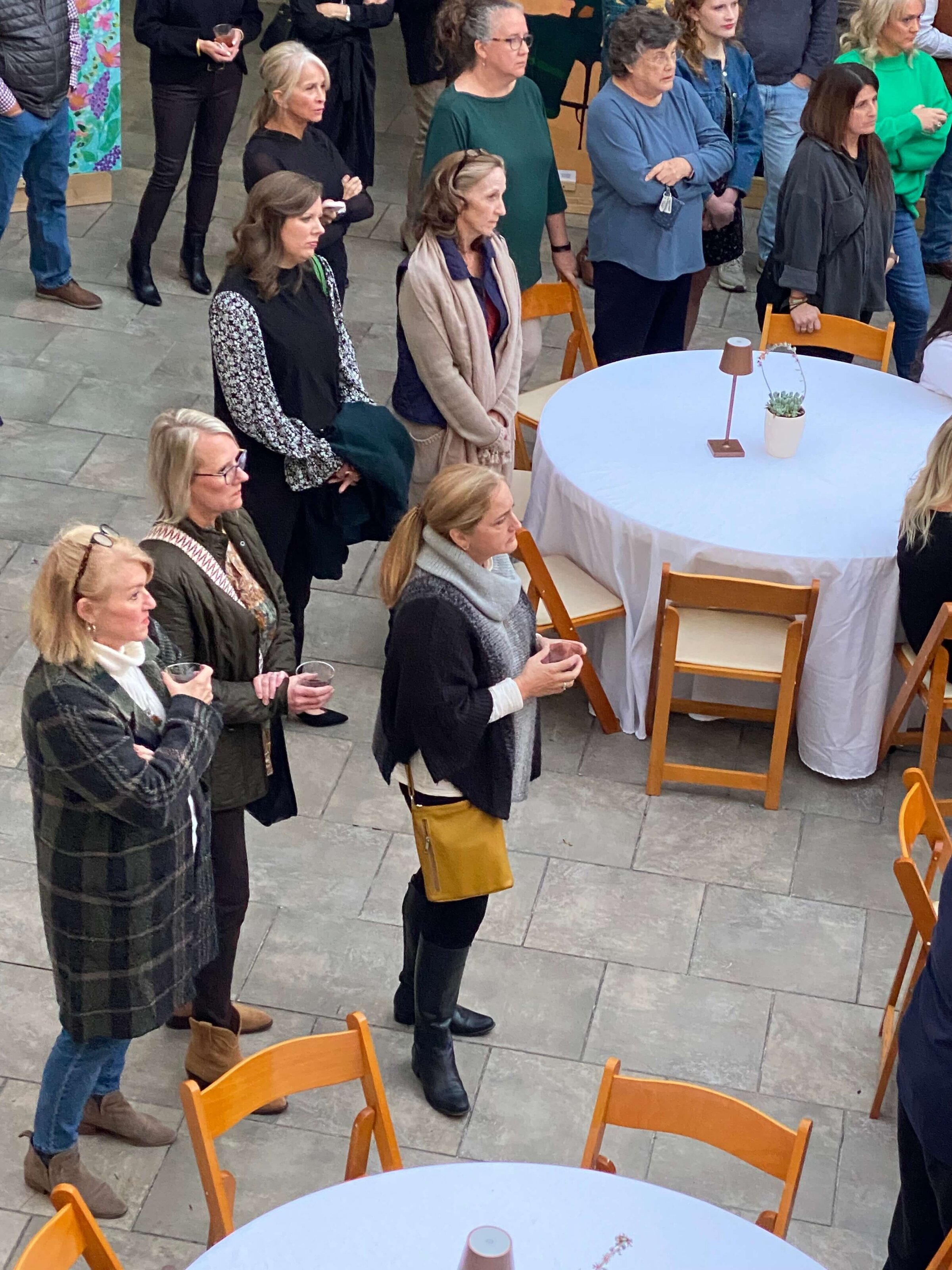 A group of people, mostly women, stand around empty round tables with wooden chairs at an outdoor event, attentively listening to something out of view. 