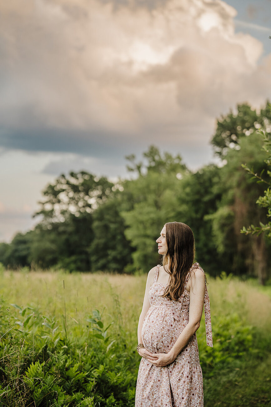woman poses in field under dramatic clouds for maternity photos