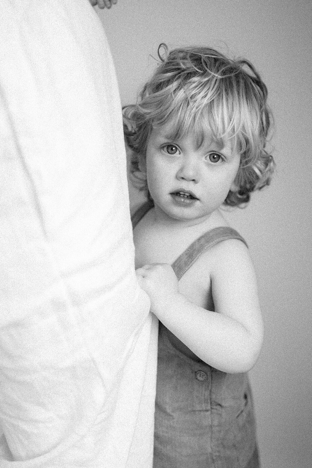 little boy in overalls holding on to mums dress