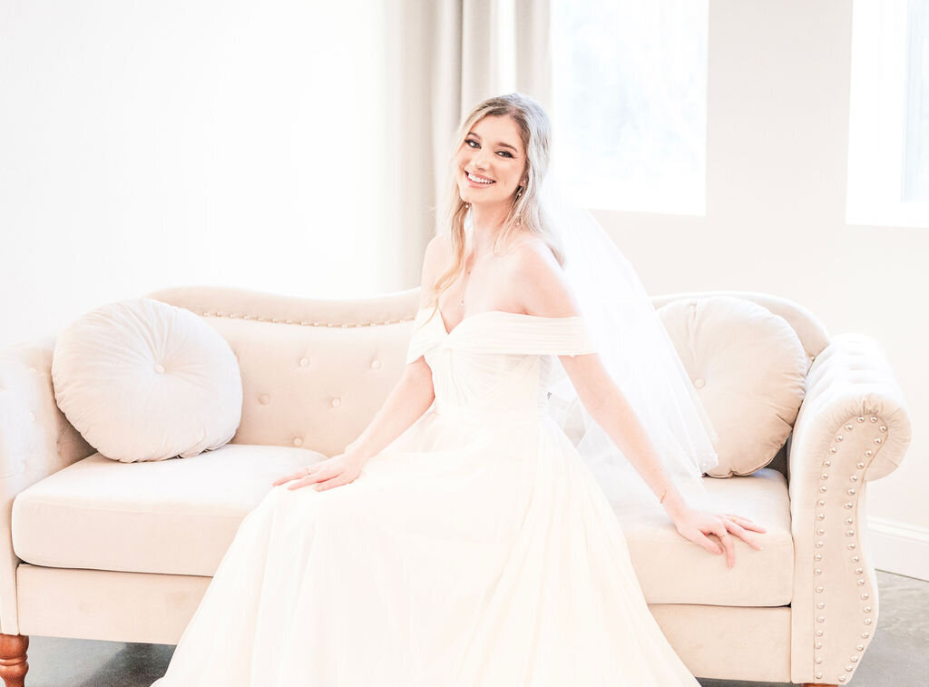 Smiling bride wearing white wedding dress sitting on sofa.