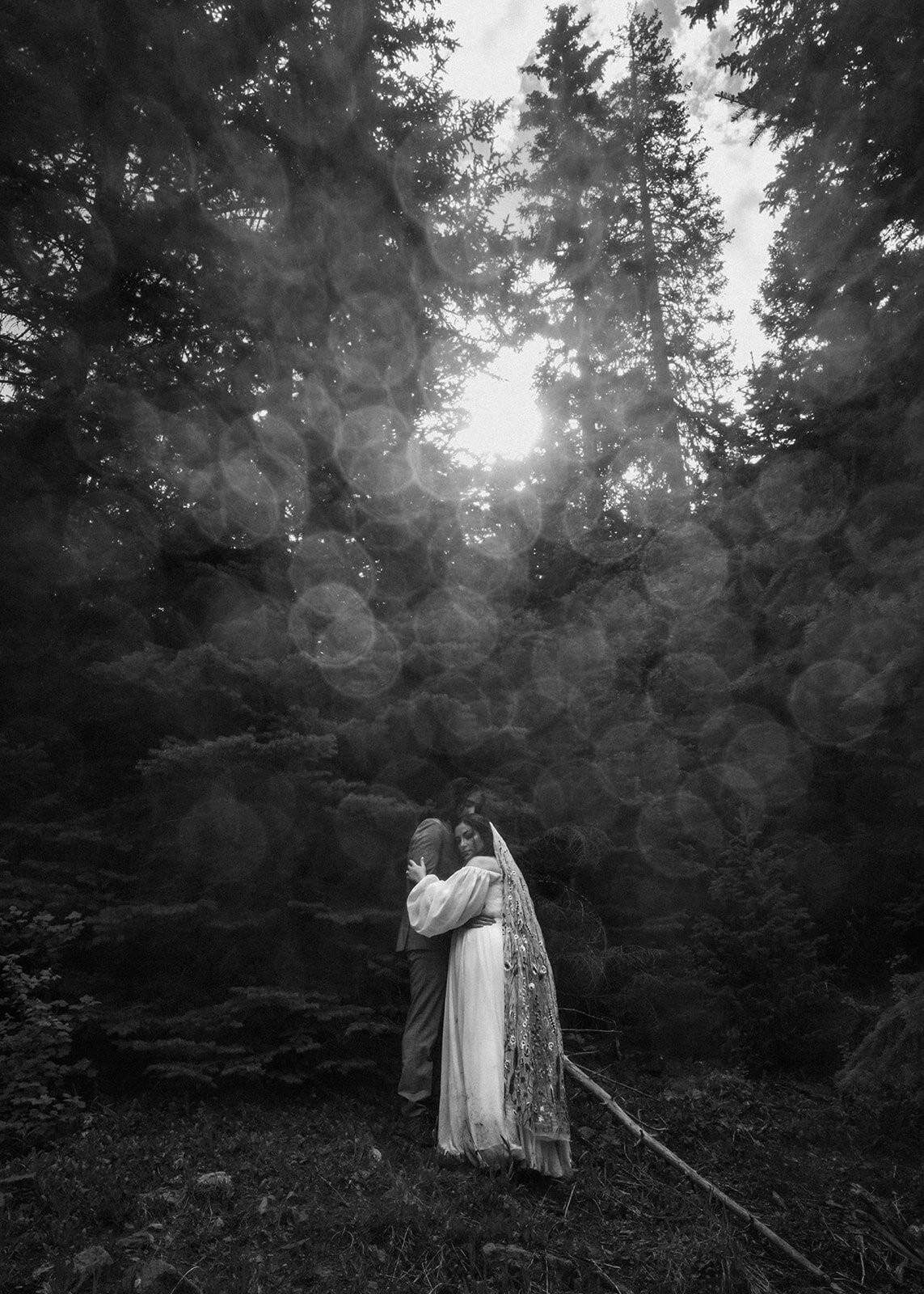 a black and white image of a bride and groom embracing under towering pines. mist covers the top of the adventure elopement photographers lens creating an almost mirror ball effect