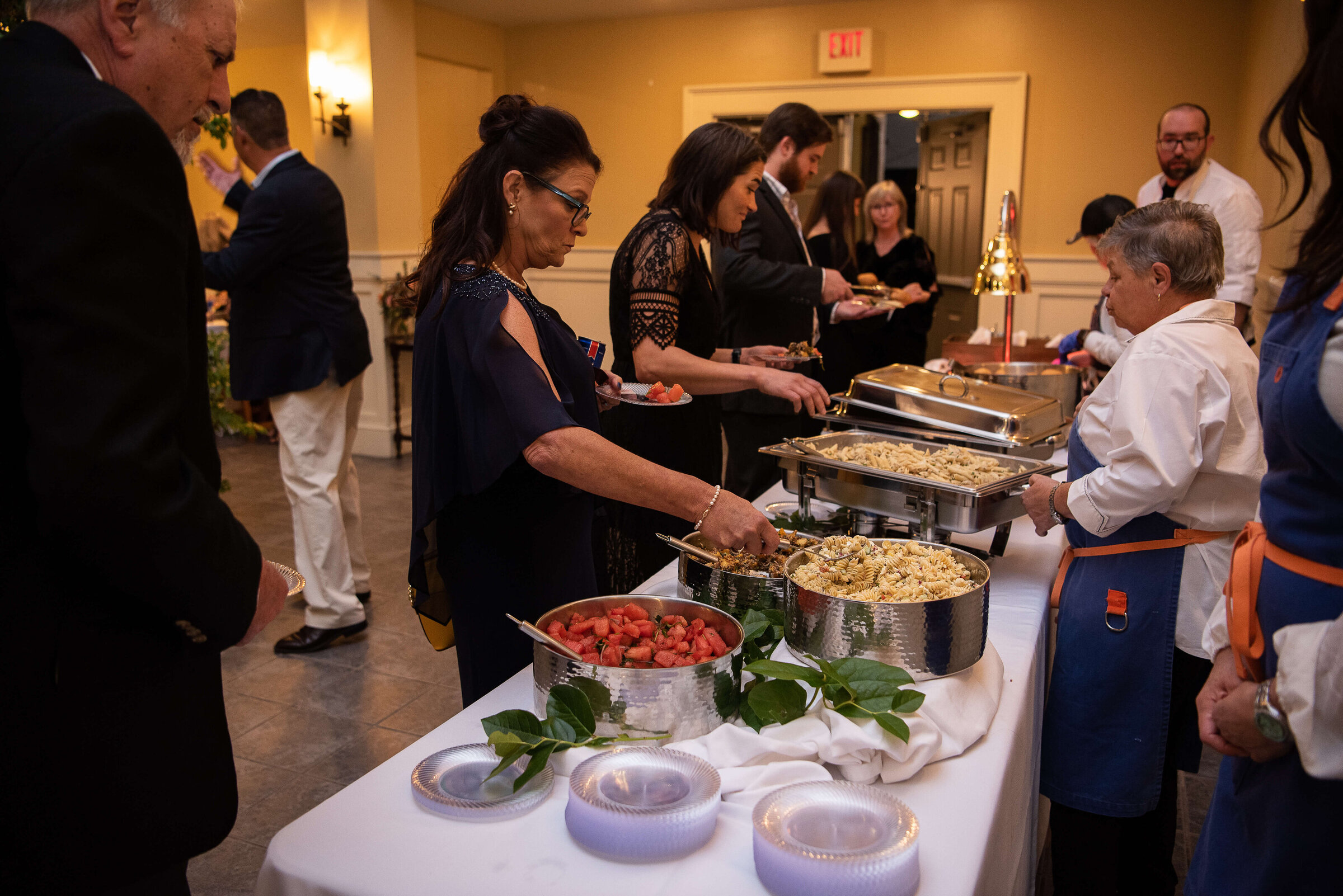 People serve themselves food from a buffet table at an indoor event, with trays of pasta and a large bowl of fruit salad.