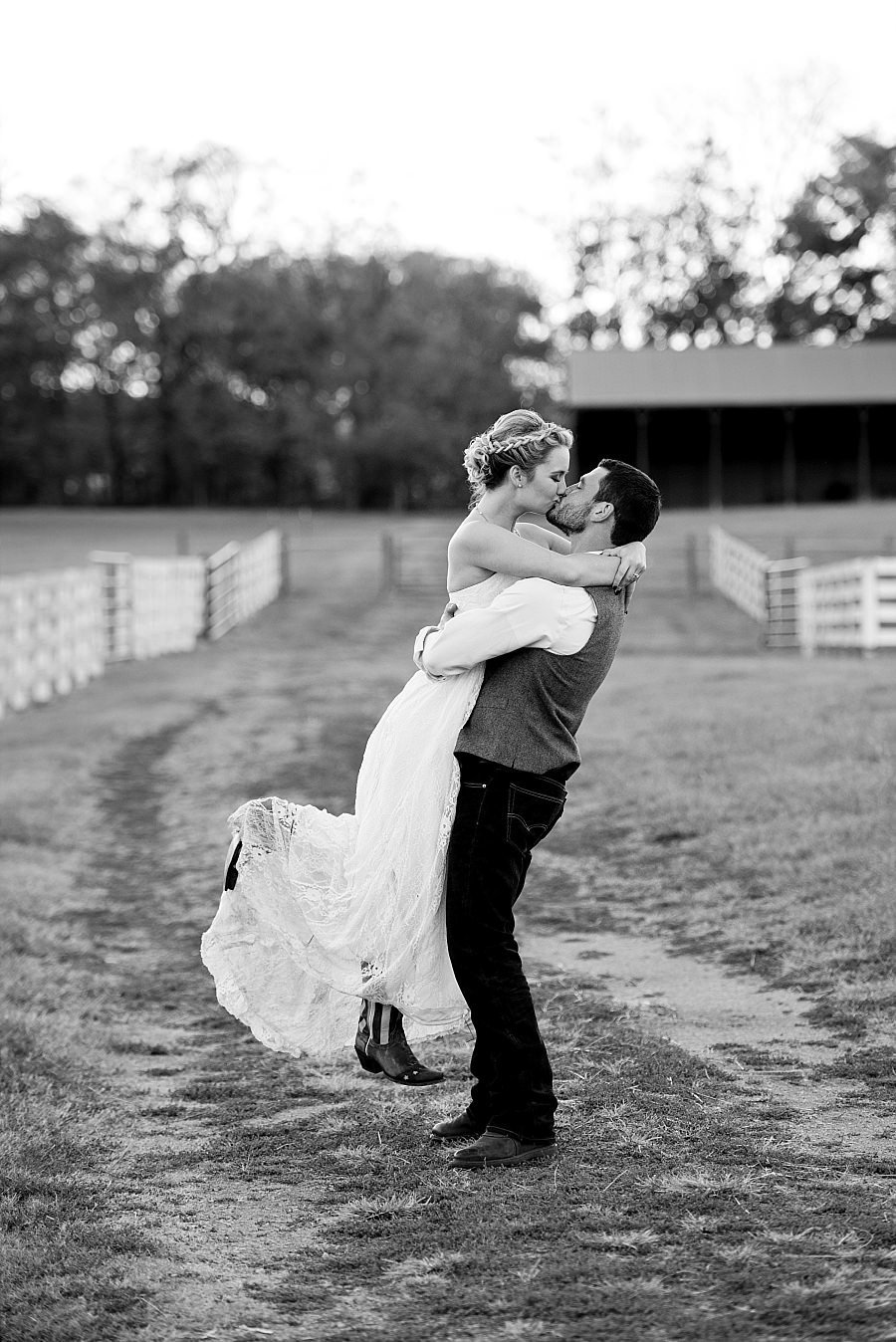 Black and White photo of groom lifting his wife and sharing a kiss on a country road with white fences around