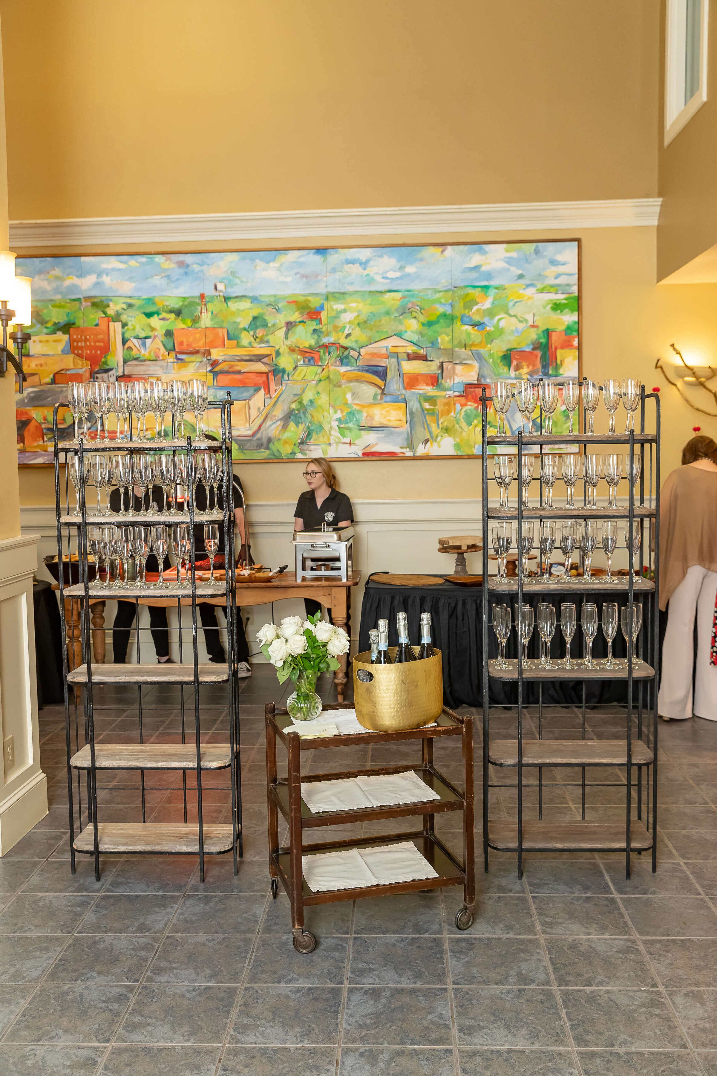 A drink station with racks of empty wine glasses, a cart with a gold ice bucket and bottles, and a person standing behind a table.