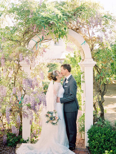 Lilac covered arch with bride in catheral veil and groom kissing in SoCal 