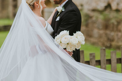 fallback shared on Invalid Date | A couple embrace, with the veil billowing around them, and a bouquet of white roses held...