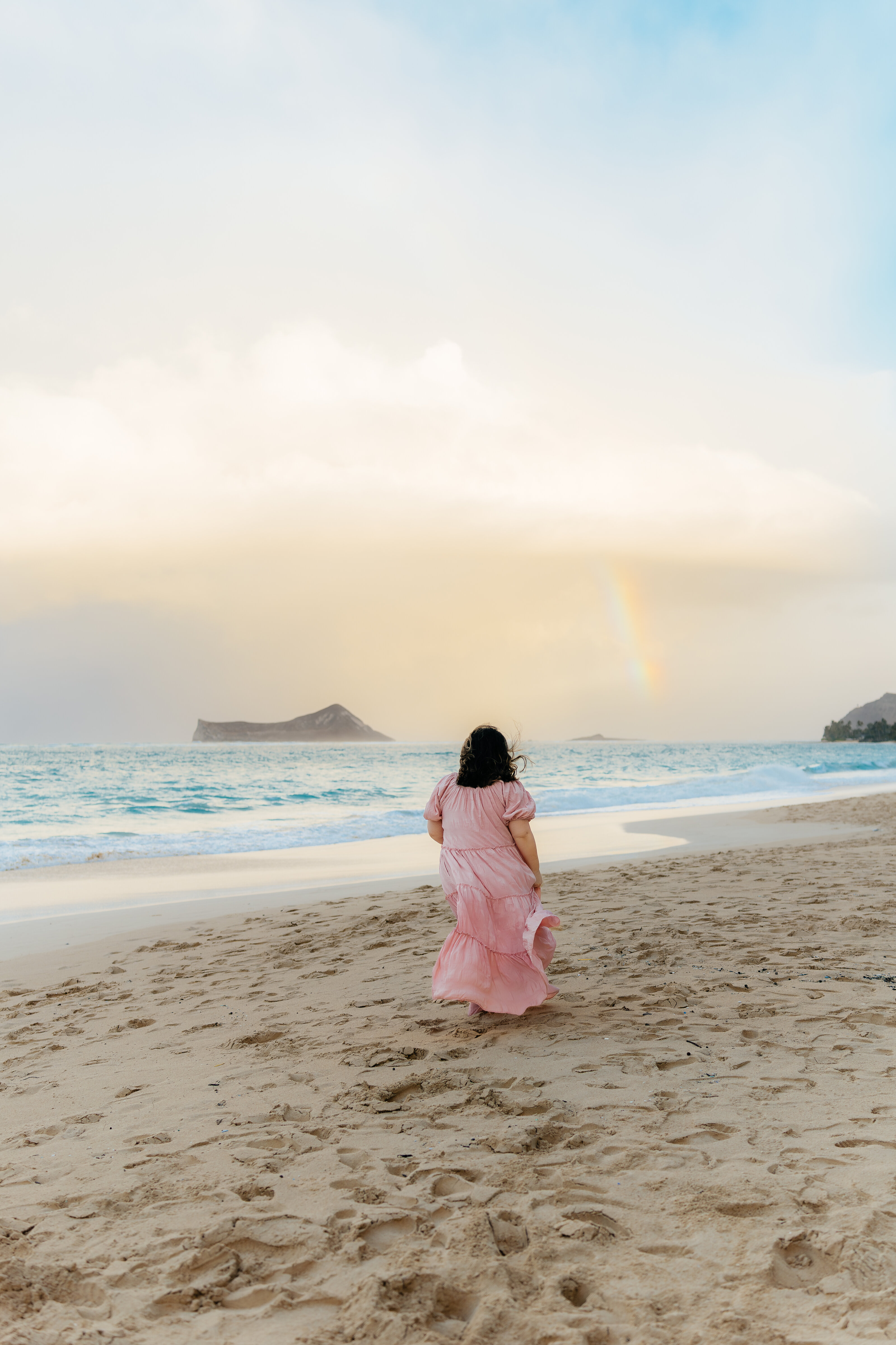 Lexi walking toward the shoreline in a pink dress as a soft rainbow appears above the ocean. Wide shot capturing the full beach scene at sunset.