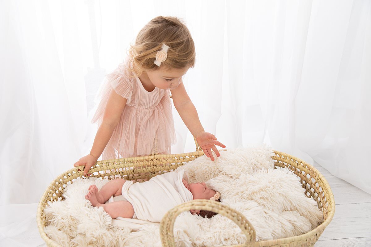 Big sister looking down at baby sister in moses basket by Baltimore Newborn Photographer