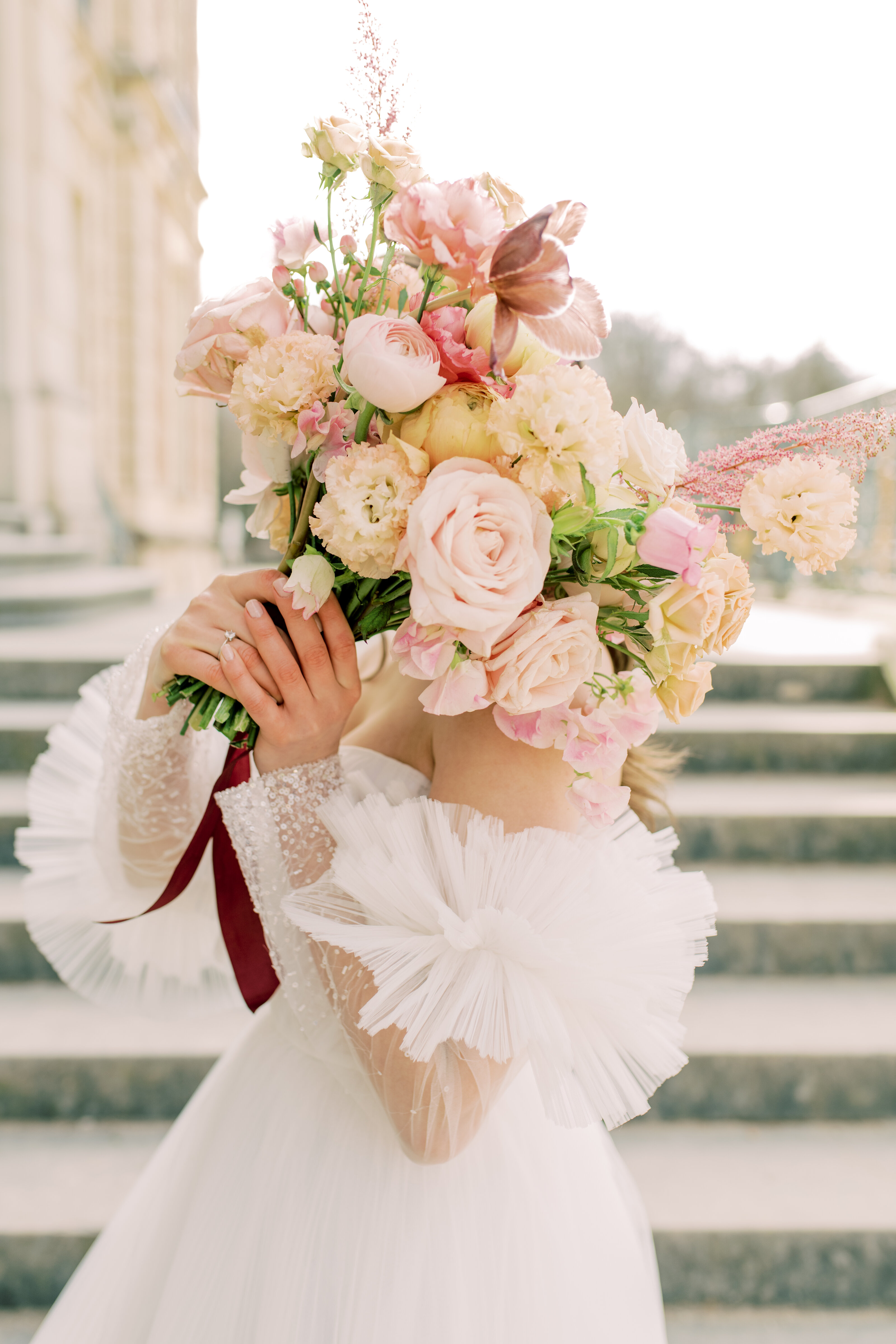 Romantic bridal portrait with the bride playfully covering her face with a pastel bouquet of roses and peonies, featuring voluminous tulle sleeves, photographed on elegant stone steps in soft natural light.