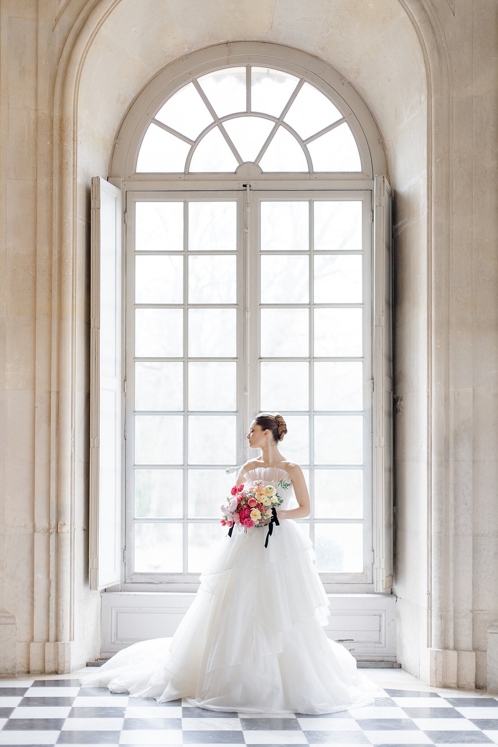 Bridal portrait in front of a large window, with a checker pattern floor. Wearing a large ballgown and holding a blush and. yellow bouquet at Chateau De Champlatreux