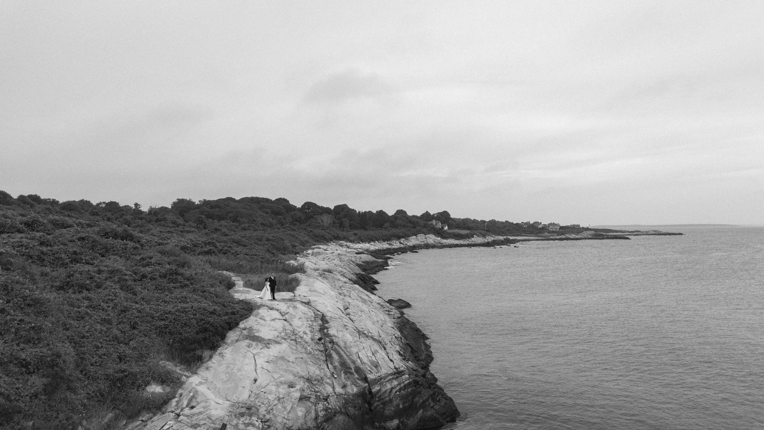 A couple stands on rocky coastline in Rhode Island, gazing at the ocean under a cloudy sky, surrounded by lush greenery.