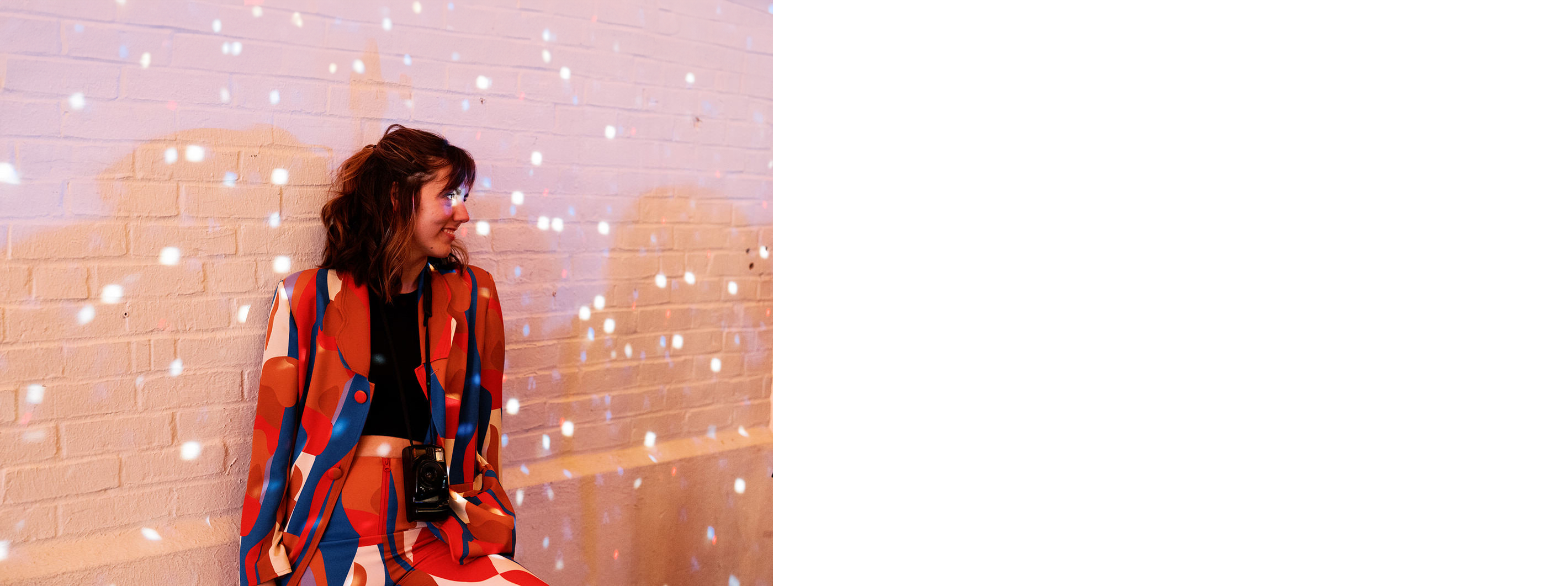 A photo of wedding film photographer Megan, who is wearing a colorful retro suit and a black shirt, she has a film camera around her neck and there is disco ball lights reflecting on her and the white brick wall behind her. She is leaning against the wall, looking to the right and smiling.