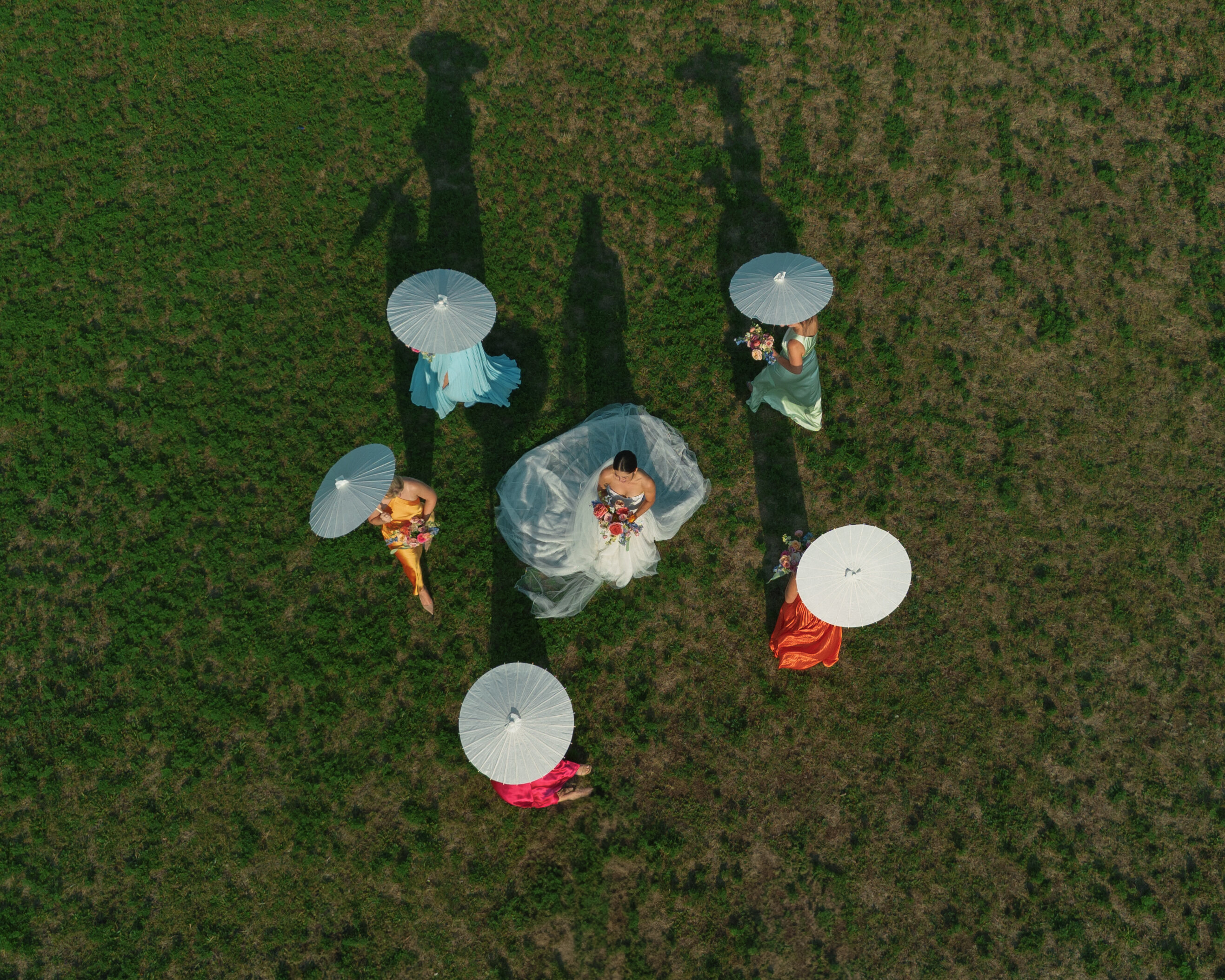 Aerial view of a bride in white surrounded by bridesmaids holding white parasols, set against a green field backdrop.