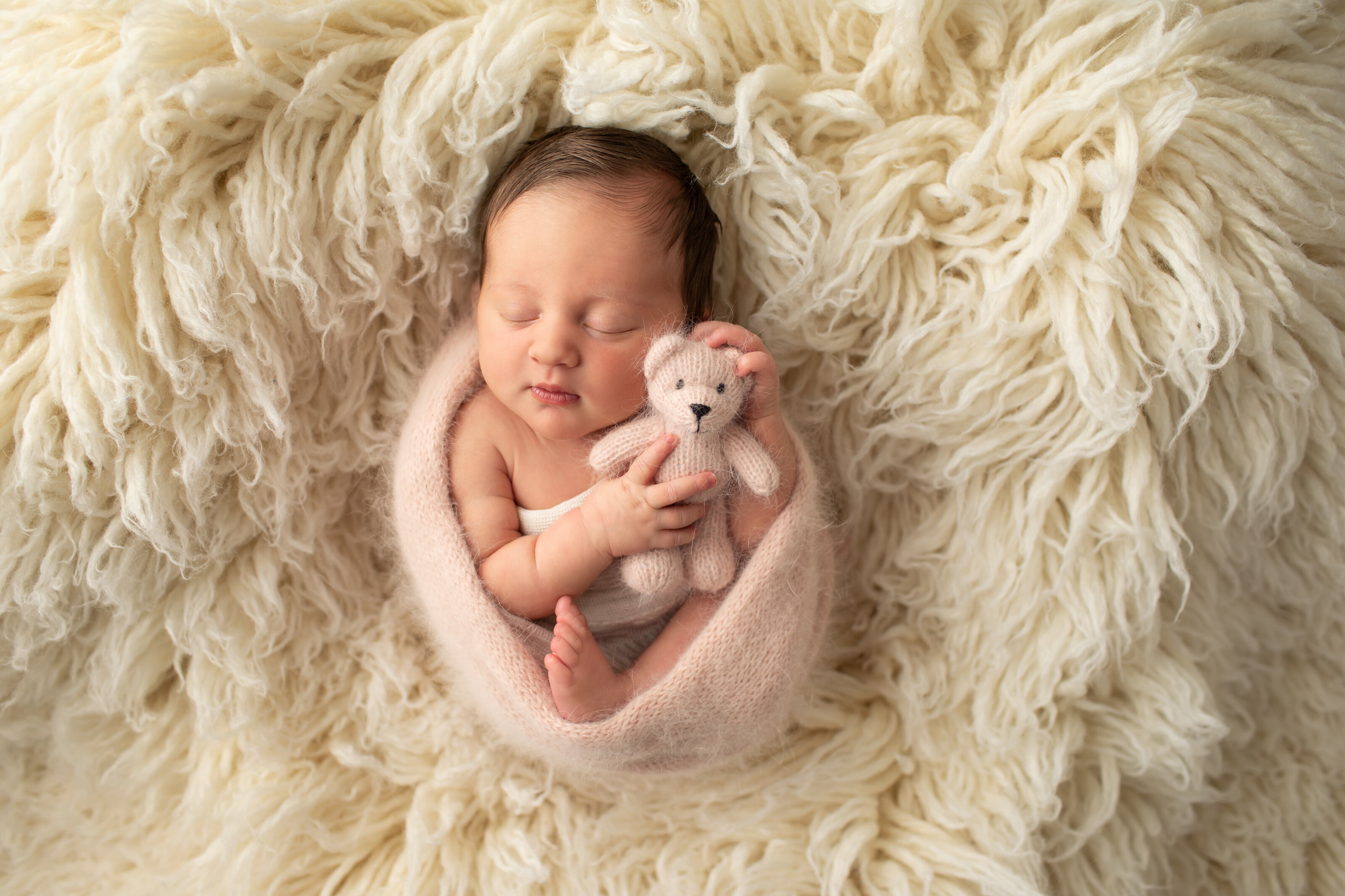 Newborn girl in pink fuzzy mohair fabric wrap and holding knit teddy bear
