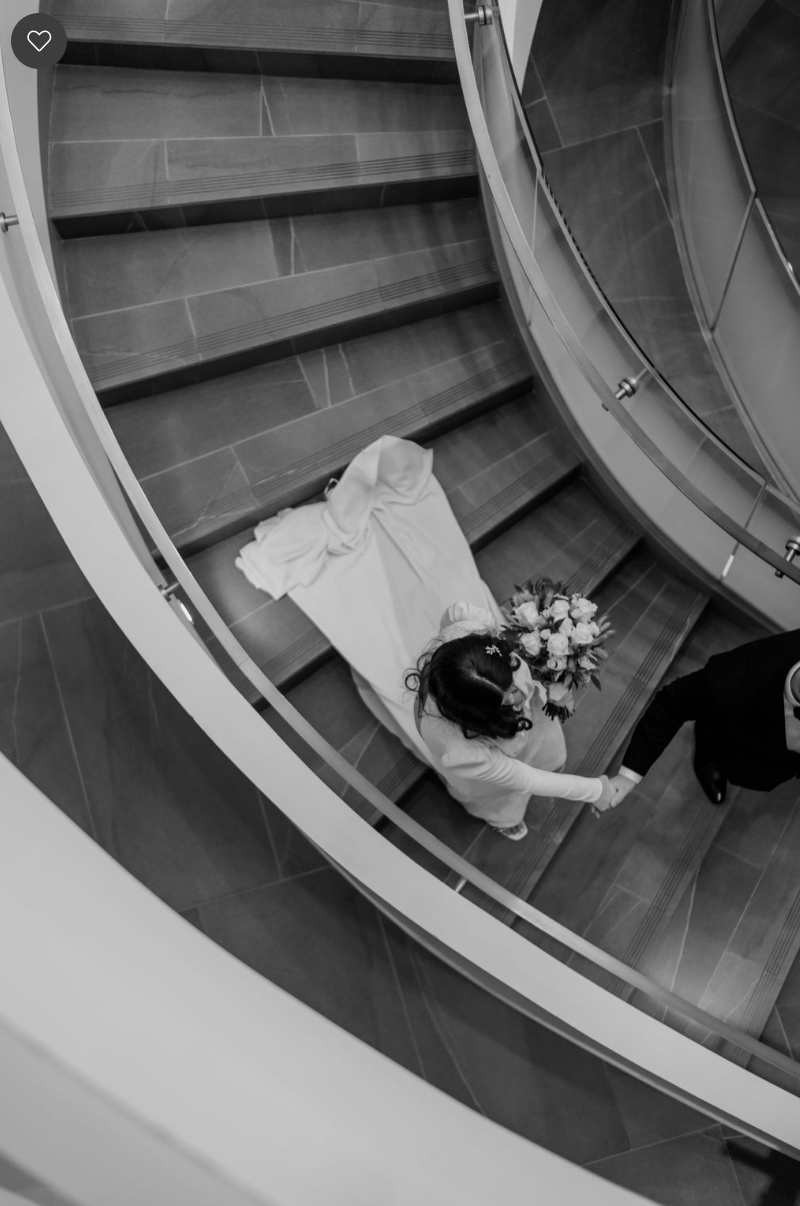 Black and white overhead shot of a bride holding a bouquet and walking hand-in-hand with her groom down a modern spiral staircase at The Pearl Hotel in Burlington, Ontario