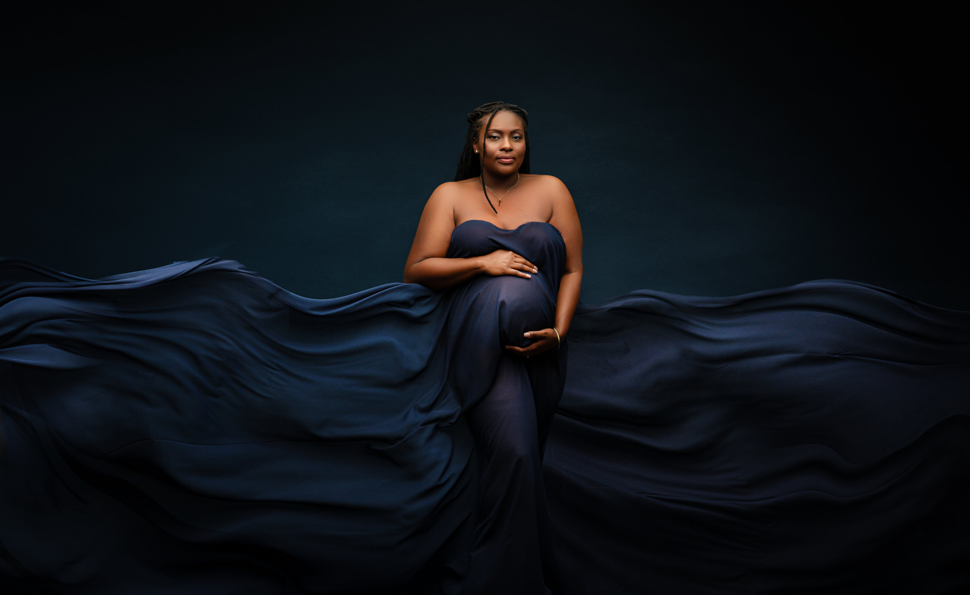 Expecting mother in soft beige floral gown with flowing train on brown backdrop, radiating confidence and grace.