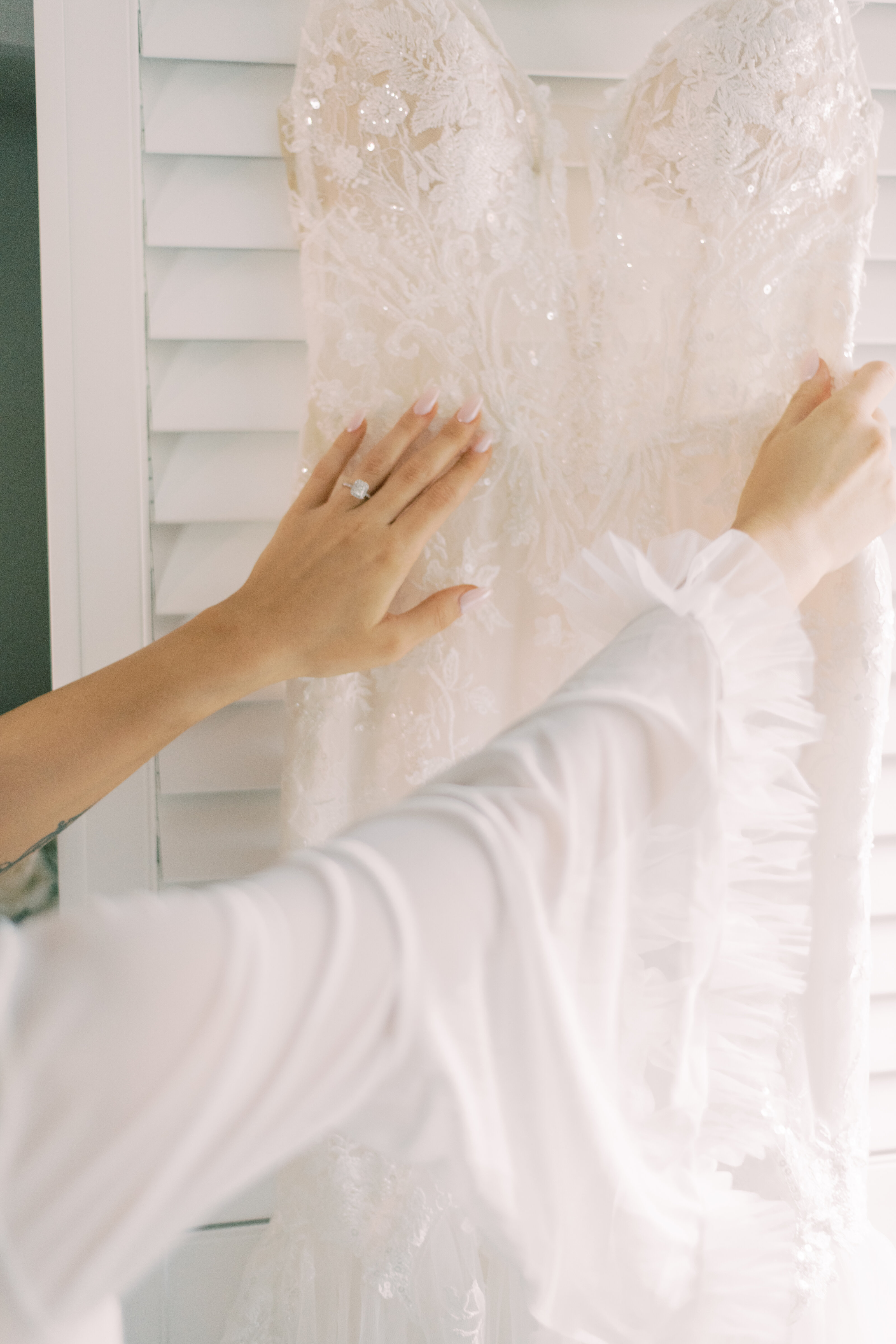 Burlington Bride’s hands adjusting a lace wedding gown on a hanger, with soft natural light and romantic ruffled sleeves in focus.