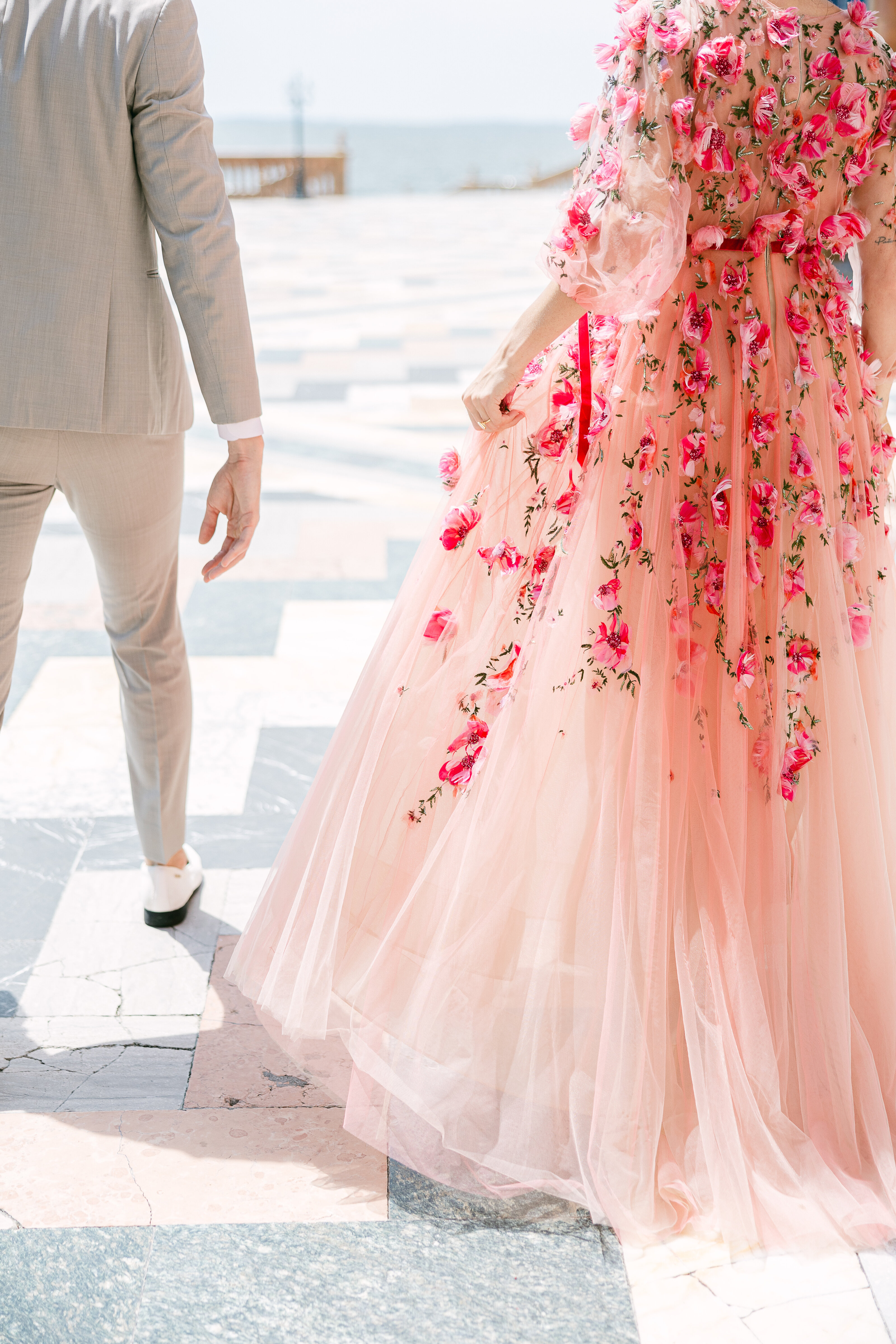 A stylish couple walks hand in hand during a dreamy engagement session in Sarasota, Florida. The bride-to-be stuns in a flowing pink gown adorned with delicate floral details.