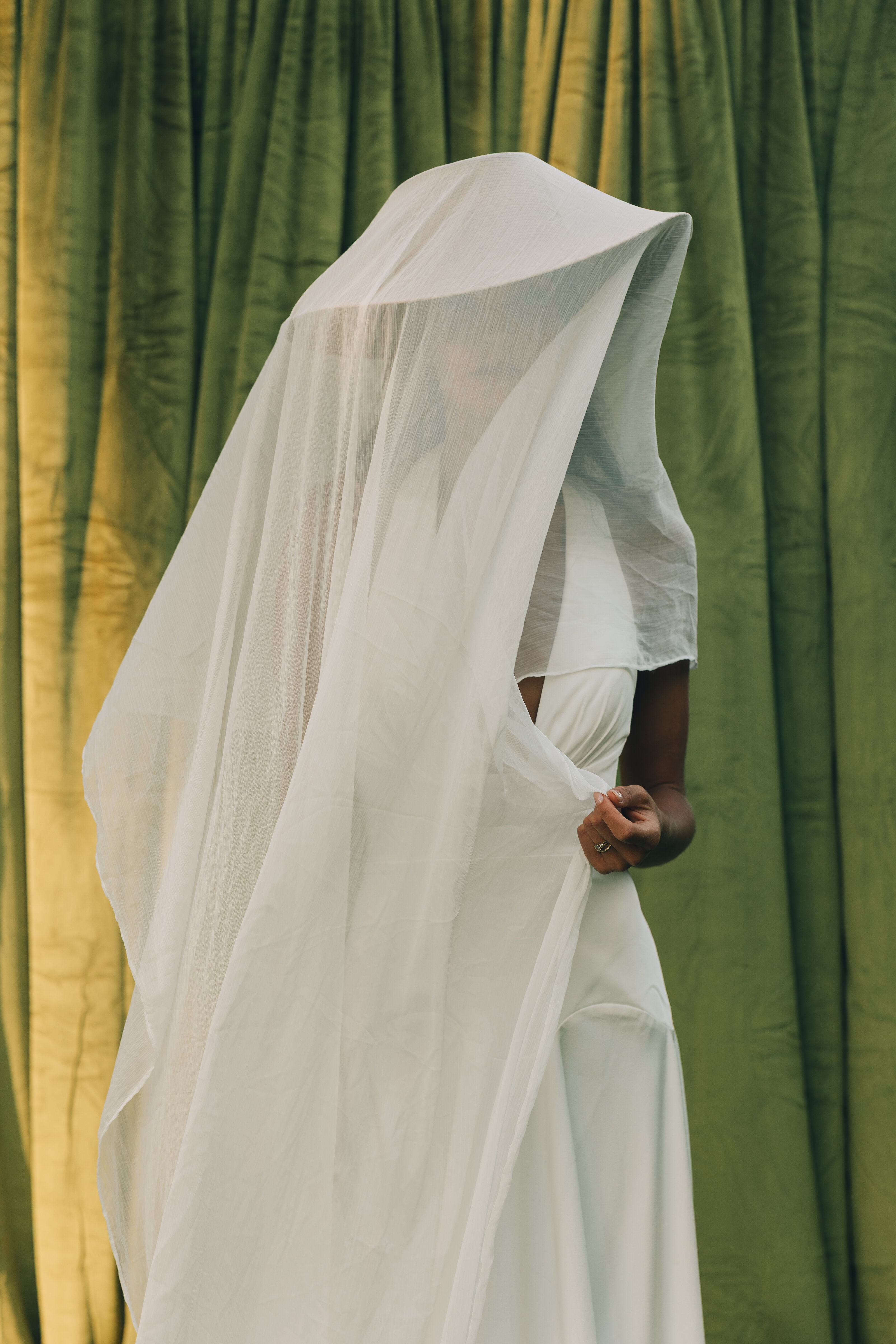 Modern bridal portrait of a woman in a minimalist white gown and oversized veil, standing against a green velvet backdrop with soft golden light.