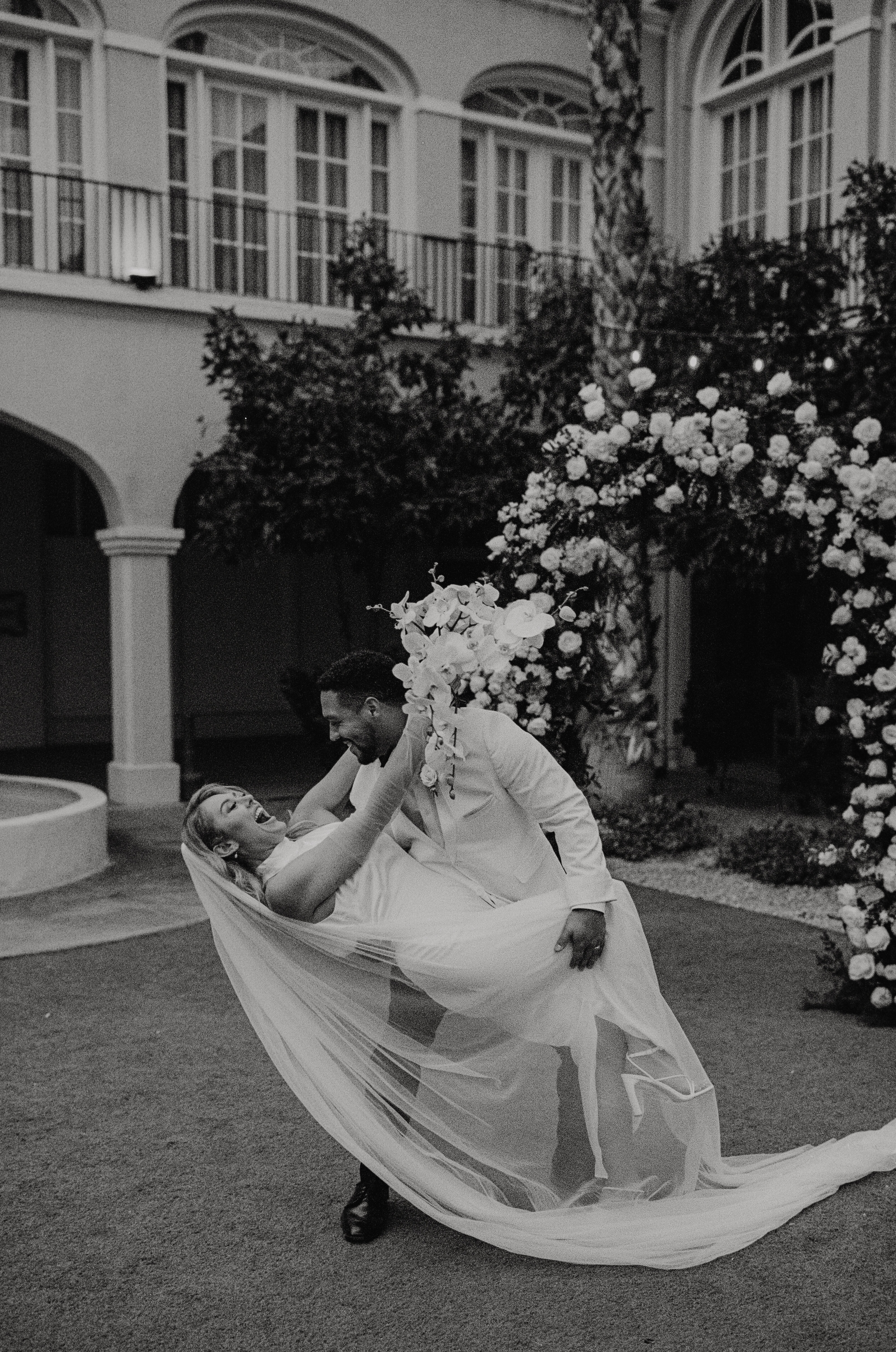 black and white photo of a groom dipping his bride while they both laugh in front of a white floral arch