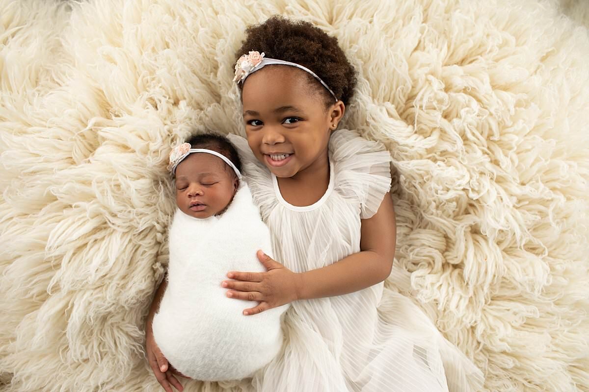 Big sister holding baby sister in cream fluffy blanket for newborn photography Columbia MD session