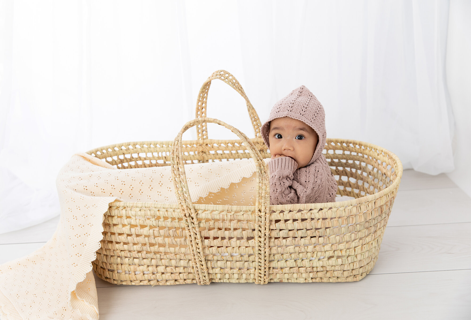 Baby girl in a mauve knit outfit and bonnet sitting a Moses basket by a white curtain