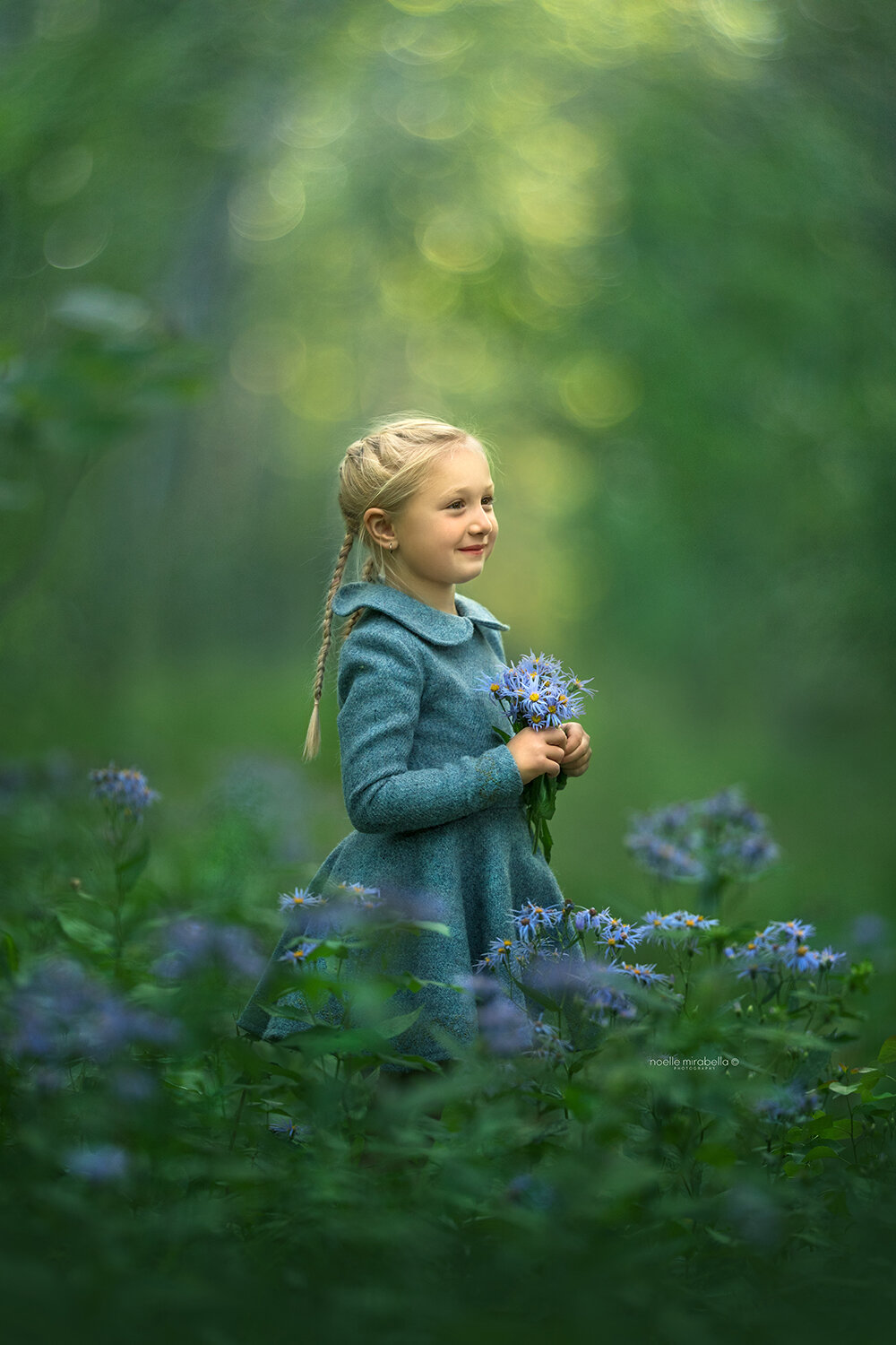 Girl in a blue wool dress holding bliue flowers, surrounded by a flower garden.