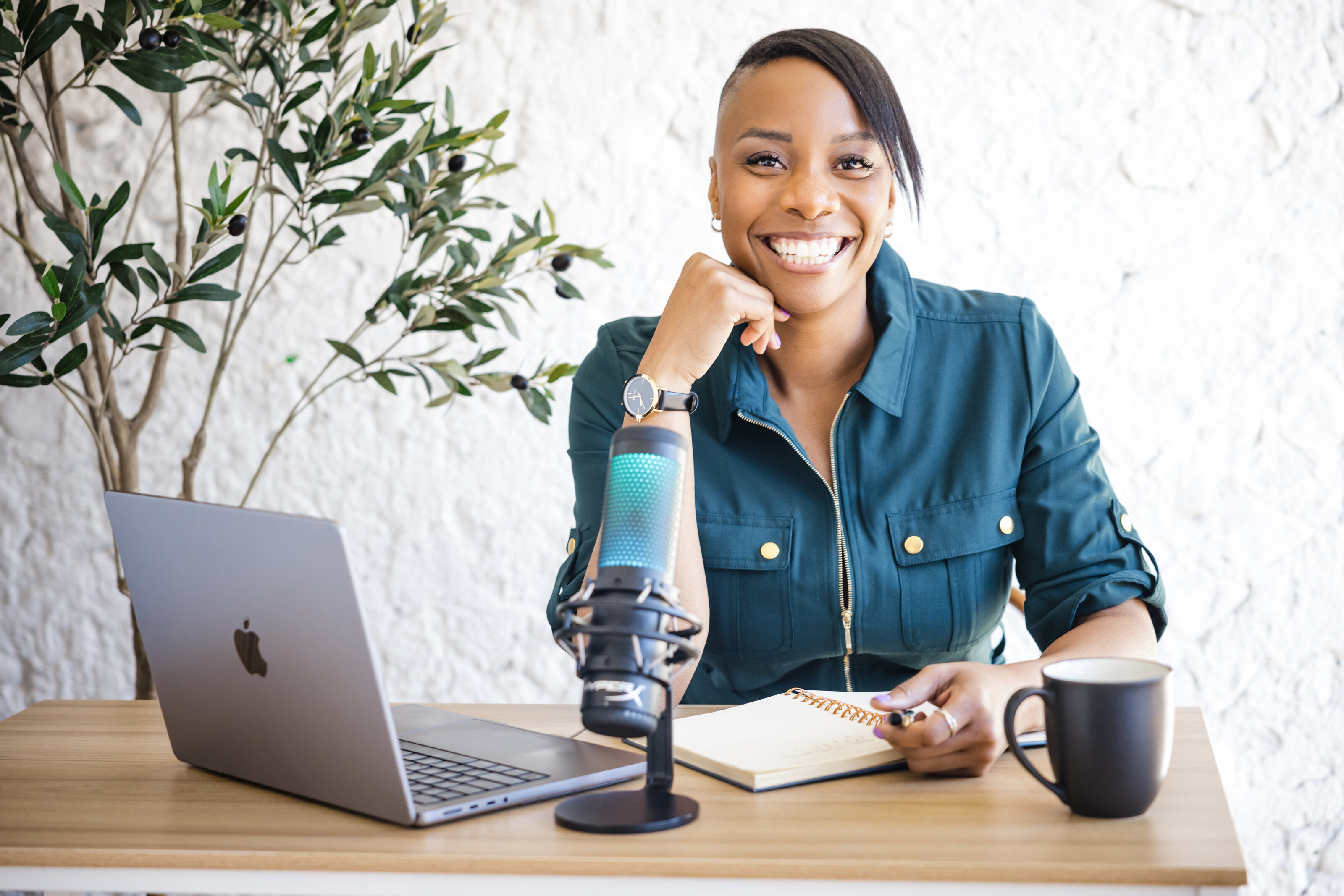 A woman sits at a desk with a laptop and a podcast microphone while laughing at the camera