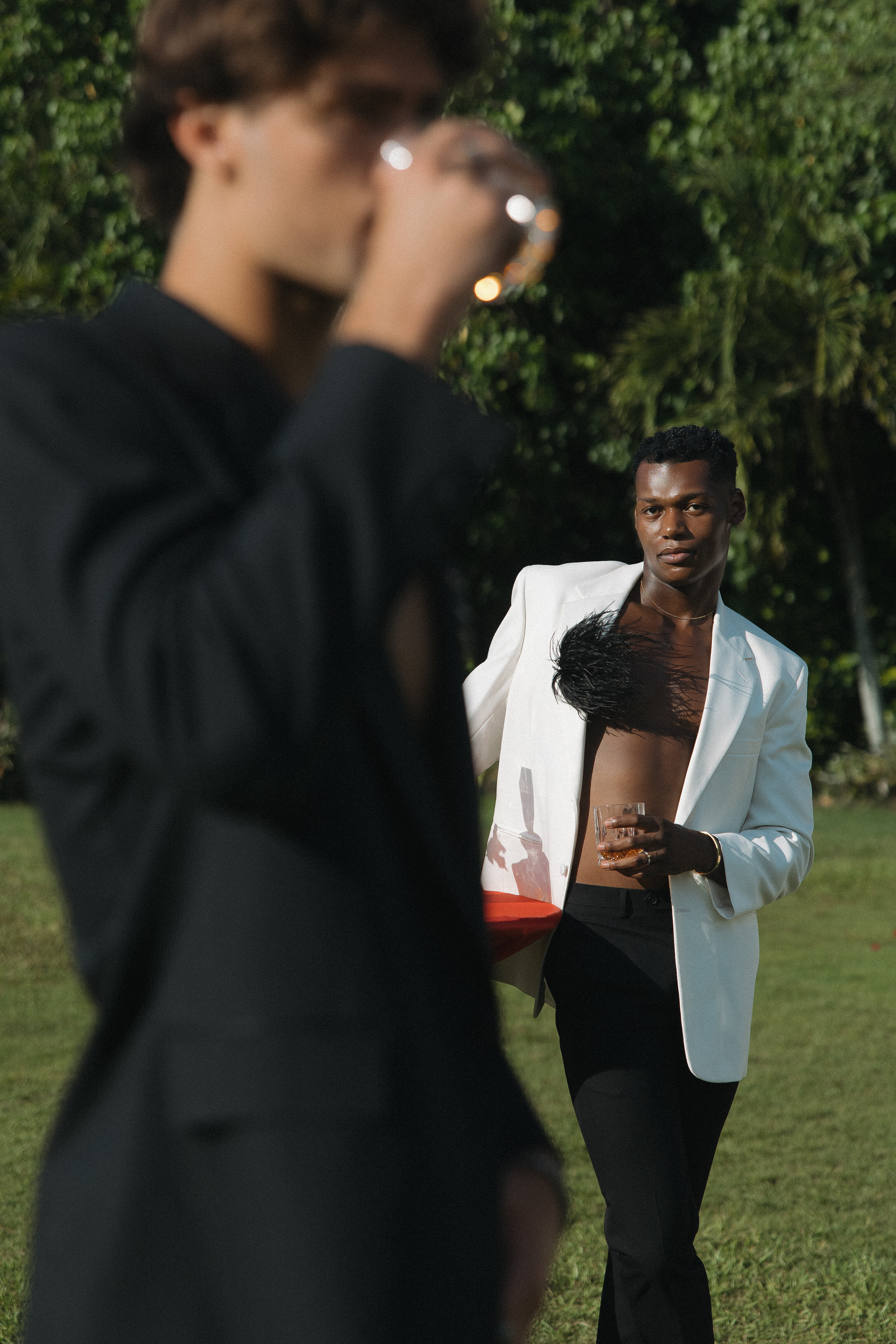 Fashion-forward outdoor portrait featuring two men in modern black and white suits, one drinking from a glass while the other poses confidently against a lush green backdrop.