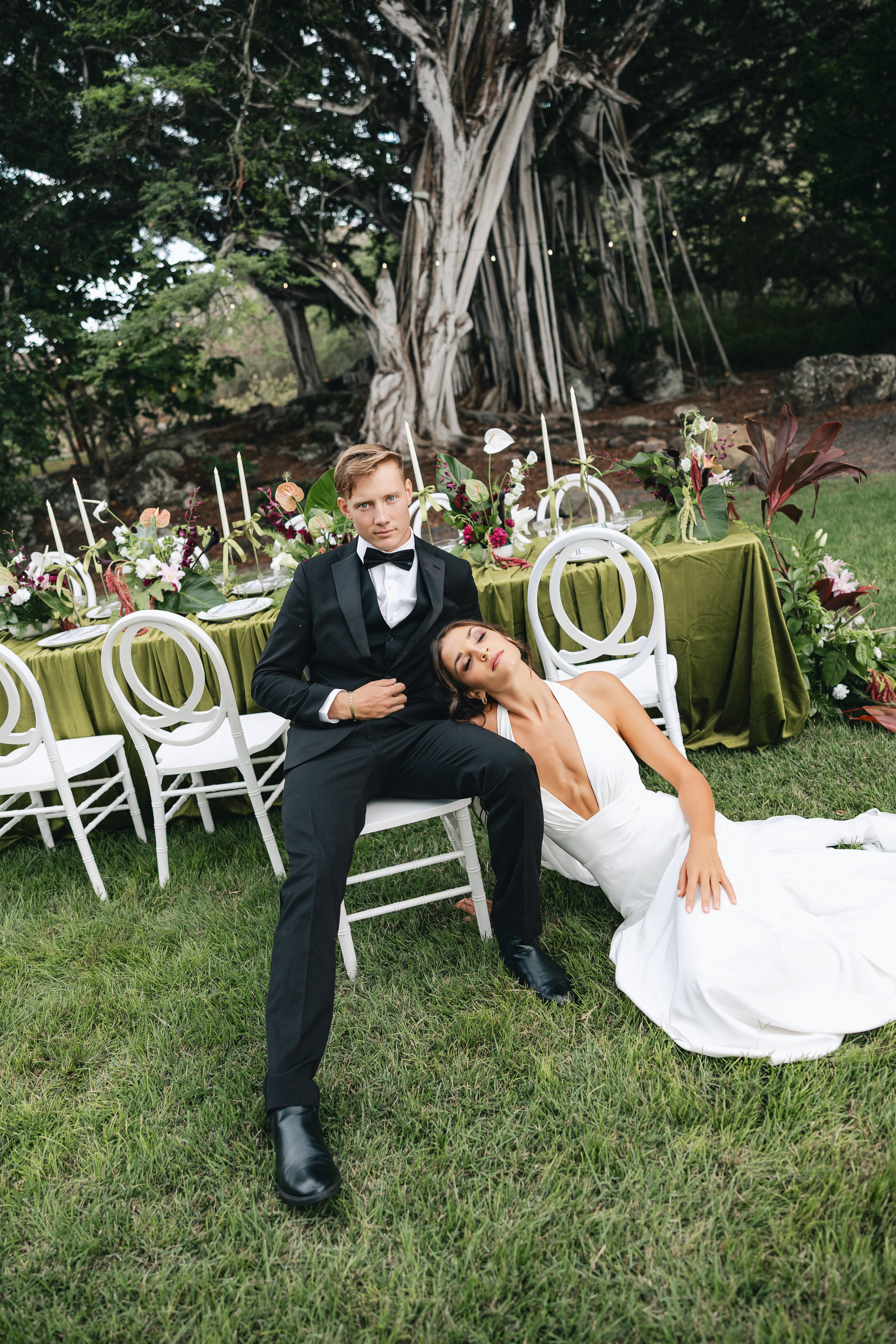 Bride in a white gown leans on the groom’s lap as they sit near an elegant outdoor reception table with green linens and floral arrangements beneath a large banyan tree.