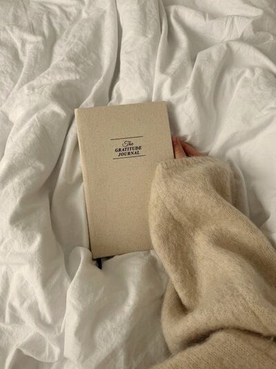 Woman sits in cozy bed wearing fuzzy beige sweater holding a gratitude journal