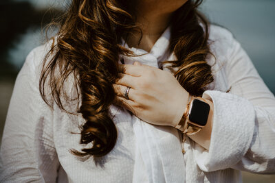 A closeup view of Anne's accessories in the background of her white dress. She is wearing her Apple Watch with rose gold bracelet, rose gold medical ID bracelet and silver wedding band and engagement band with stones.