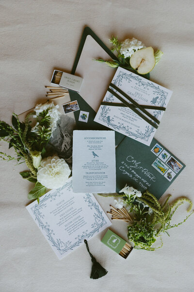 Flatlay of a custom wedding invitation suite featuring florals, envelopes, matchbooks and pears representing full service design and calligraphy by Life in Letters.