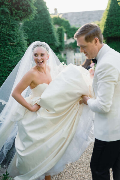 groom helping a bride walk through euridge manor luxury wedding venue gardens by holding her dress as bride laughs