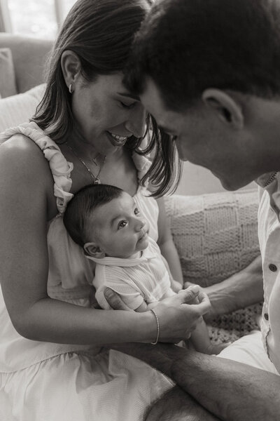 Black and white image of parents leaning in close, gazing at baby on their lap.