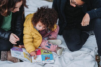 Curly haired toddler girl reading a book with her two mums in Hampstead Heath London