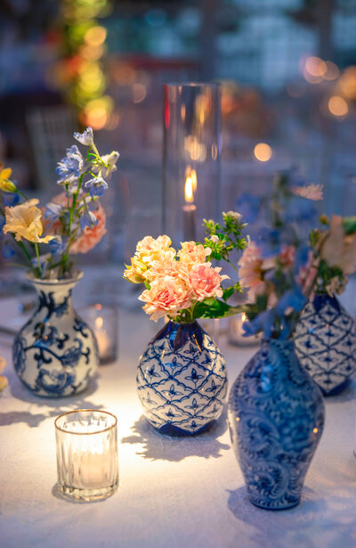 A table adorned with blue and white patterned vases holding pink and yellow flowers. Soft candlelight illuminates the scene, enhancing the elegant and serene atmosphere.