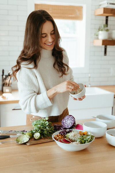 Woman with brown hair and a tan sweater seasoning food in the kitchen