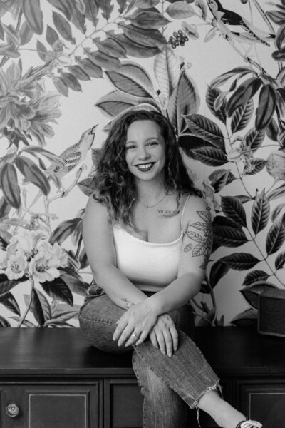 A smiling woman with curly hair, wearing a white tank top and jeans, sits on a bench in front of vibrant, leafy wallpaper with bird illustrations. This black-and-white image perfectly captures the creative spirit of a white label designer.