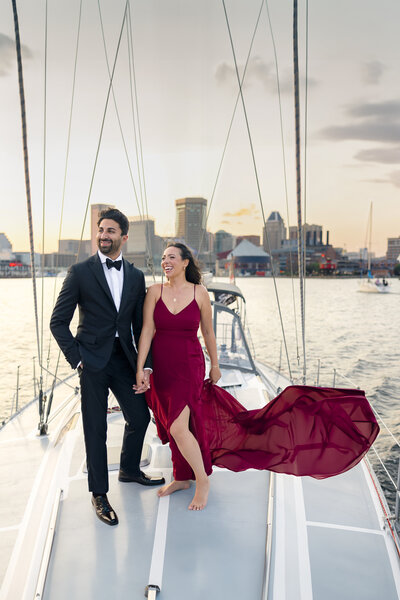 A couple stands on a sailboat at sunset, with city buildings in the background. The man is in a black tuxedo and the woman wears a flowing burgundy dress. They hold hands and smile, enjoying the scenic view.