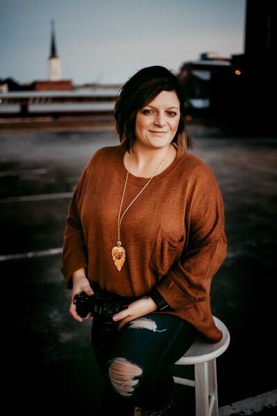 Person sitting on a stool outdoors, holding a camera, wearing a brown sweater and ripped jeans. A church steeple is visible in the background.