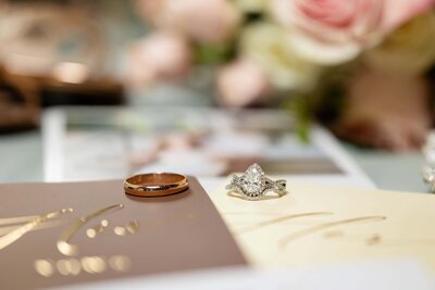 A close-up flat lay of a gold wedding band and a pear-shaped diamond engagement ring placed atop elegant wedding invitations. Soft pink roses and blurred photos in the background add a romantic, thoughtful touch to the scene. Captured by Heartful Films, a Jacksonville wedding photographer and videographer who highlights sentimental details and luxury storytelling in every celebration.