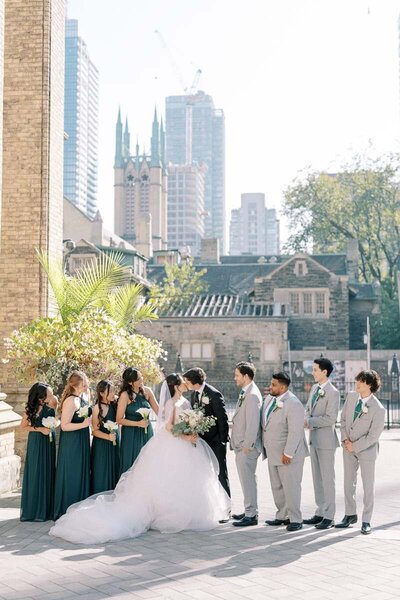 Bride and groom kissing surrounded by wedding party, cityscape in the background, joyful moment in Toronto