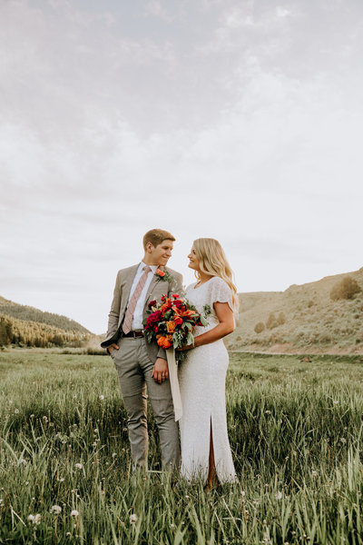 photo of bride and groom looking at each other and holding a colorful bouquet in the valley of mountains in Idaho during bridal session, photo by Kaitlyn Neeley