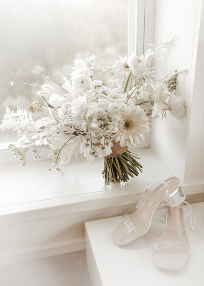 White and green bouquet leaning on white window sill in front of a pair of heels