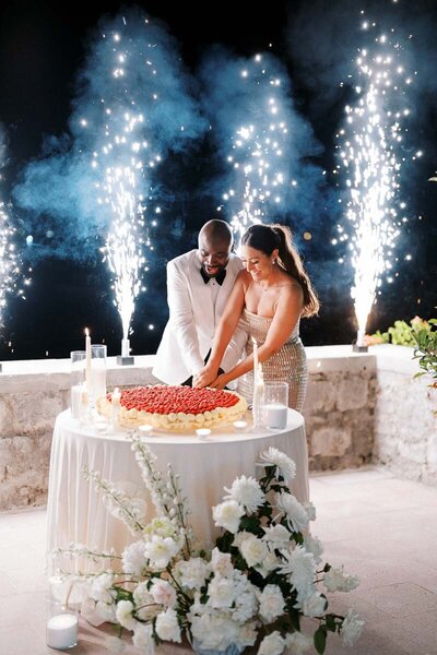 Bride and groom cutting heart-shaped fruit cake during wedding night celebration with fireworks