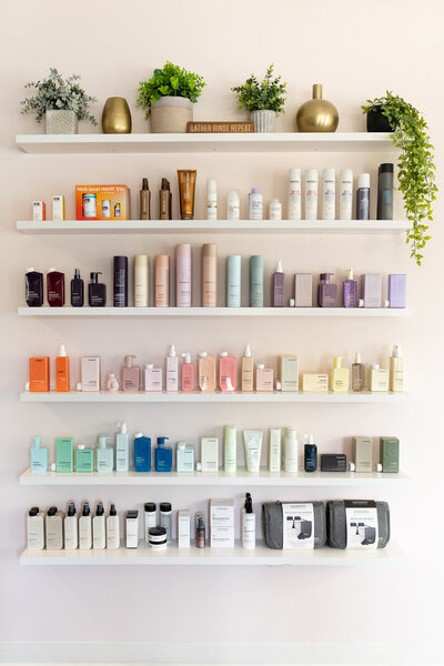 Wall of salon products with green plant decor on top shelves of salon wall. 