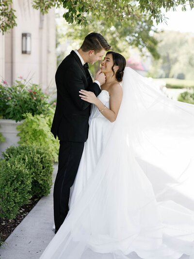 Bride and groom share a romantic kiss outside under natural sunlight, veil flowing in the breeze.